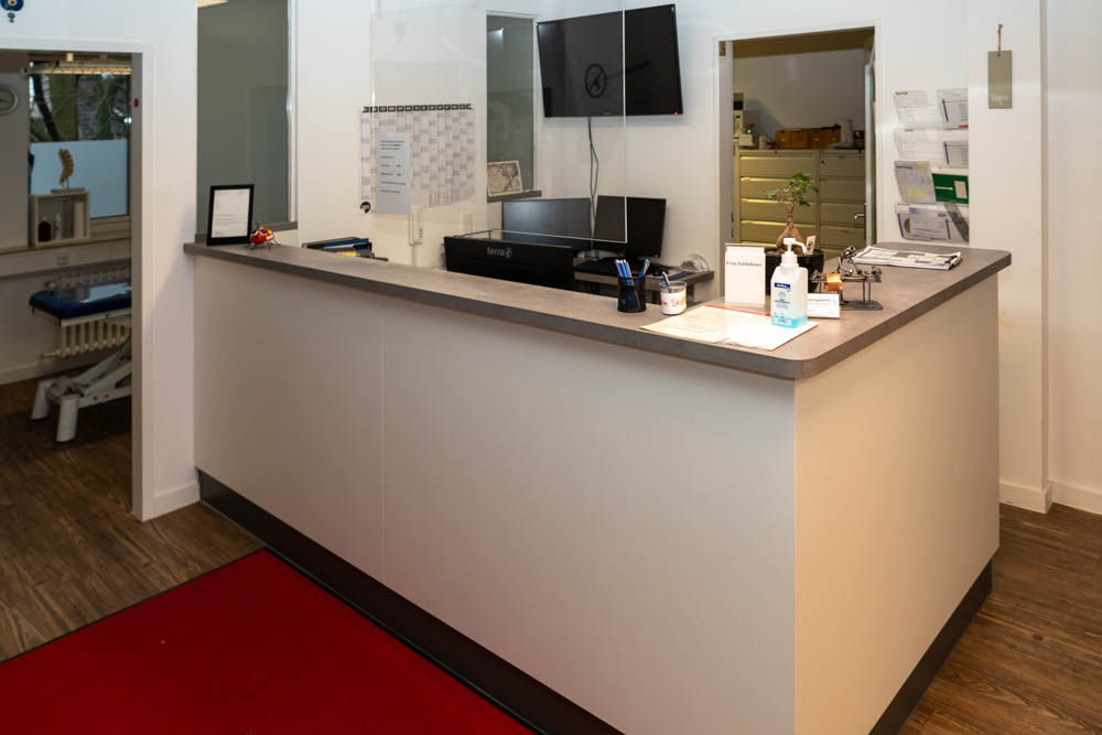 Empfangstheke in einer Physiotherapiepraxis, mit modernen Möbeln und einem Schreibtisch, auf dem mehrere Arbeitsutensilien liegen.
